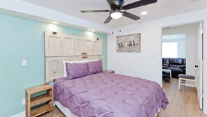 Ein Schlafzimmer mit Bett, Deckenventilator, Holz-Kopfteil, einer Bank und einer Tür zu einem anderen Raum.