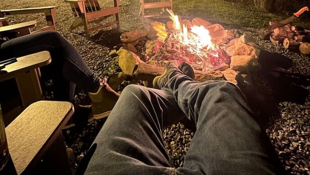Une personne se détend près d’un feu de camp, assise sur une chaise, avec un barbecue à proximité.