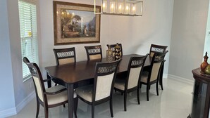 Uma sala de jantar com uma mesa e cadeiras de madeira escura, um candelabro, um quadro emoldurado e uma janela com persianas.