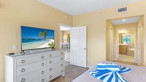 Ein Hotelzimmer mit Bett, Kommode, Fernseher, der eine Strandansicht zeigt, und einem durchgehenden Türspalt einsehbarer Badezimmers.