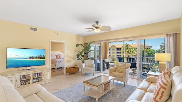 Ein geräumiges Wohnzimmer mit großem Flachbildfernseher, bequemen Sitzgelegenheiten, Deckenventilator und Blick auf den Außenbalkon.