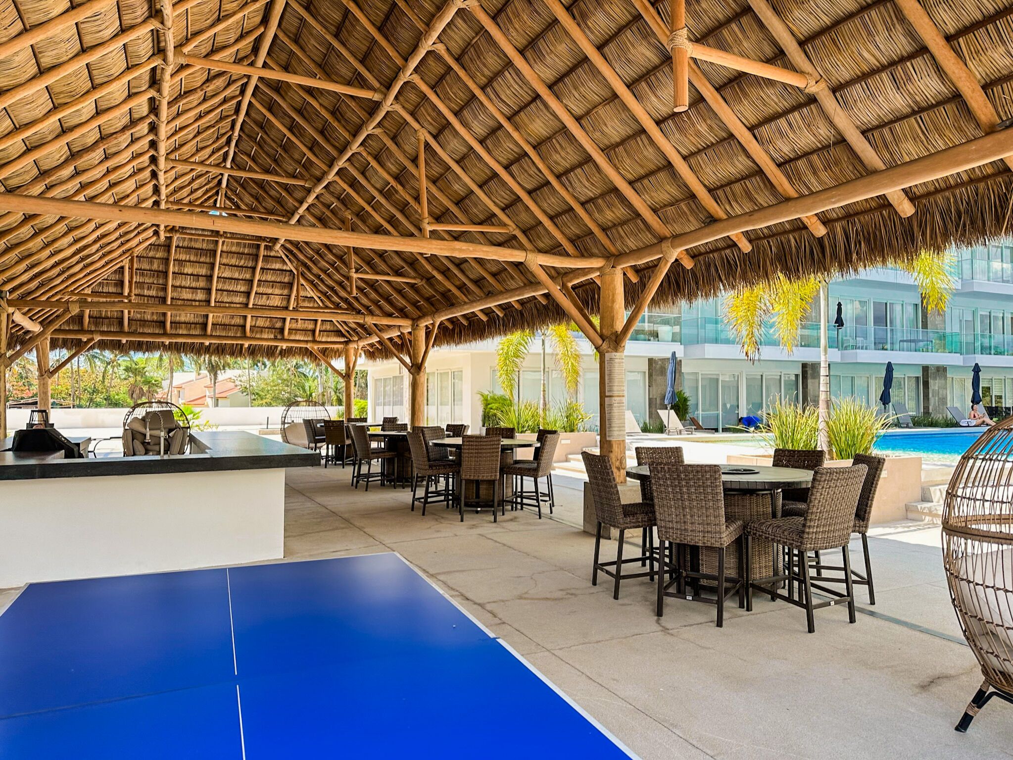 An outdoor seating area with tables and chairs, a pool, and a thatched roof structure.