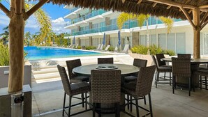Un área junto a la piscina con techo de paja, mobiliario de mimbre y vista a un edificio de varios pisos con balcones.