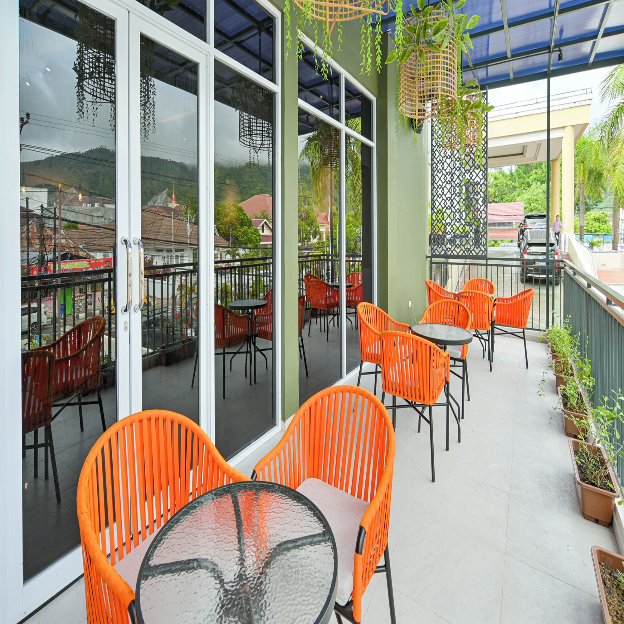 An outdoor patio with orange chairs and tables, a glass door leading to a view of buildings and hills, and hanging plants.