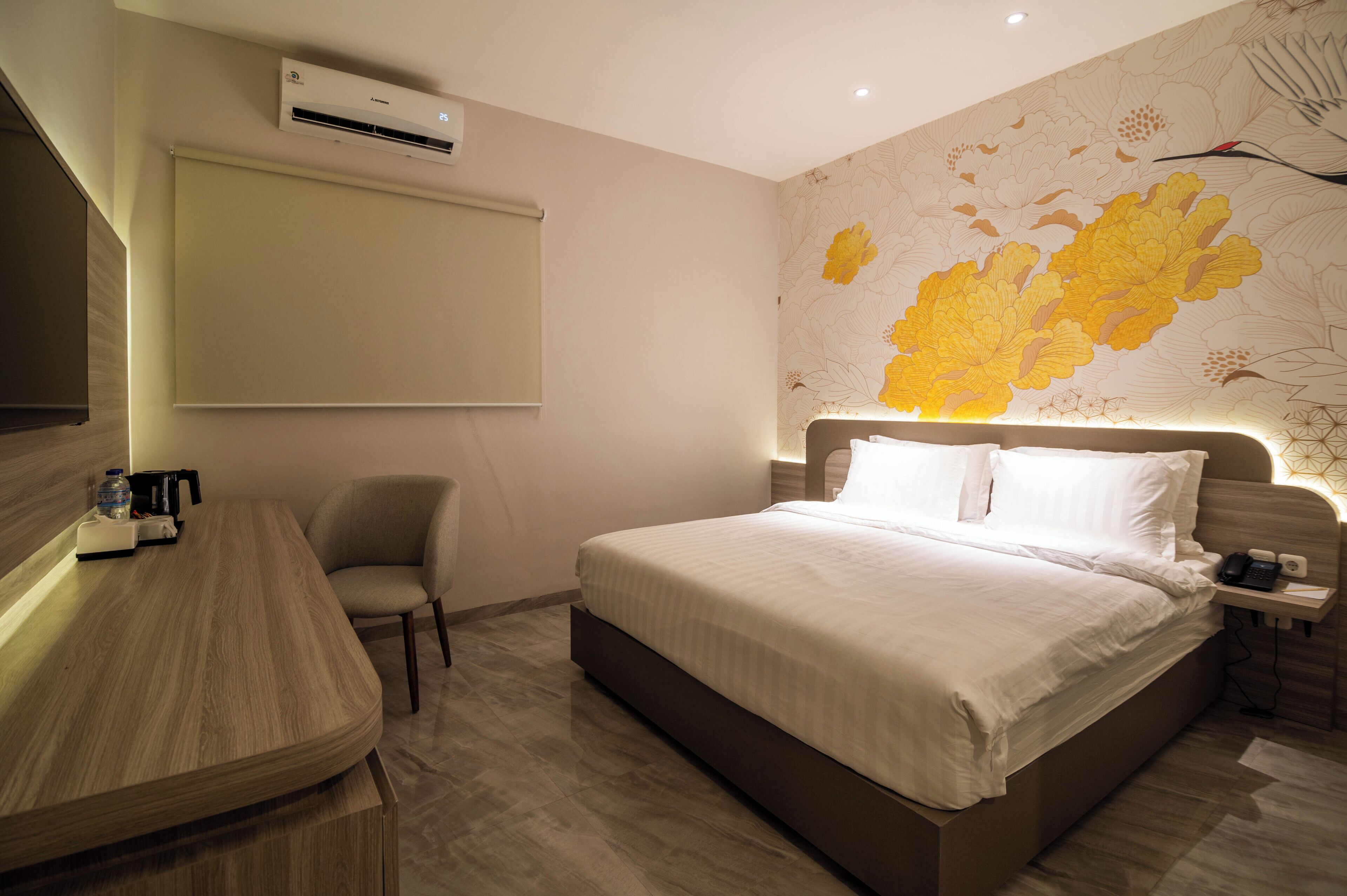 A modern hotel room with a large bed, a desk with a chair, a bedside table with a phone, and a wall-mounted air conditioner.