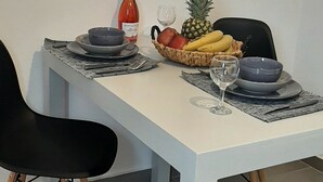 Ruokapöytä, jonka päällä on valkoinen pöytäliina, mustat tuolit, viinipullo, ananas, banaaneja ja kulho täynnä hedelmiä.