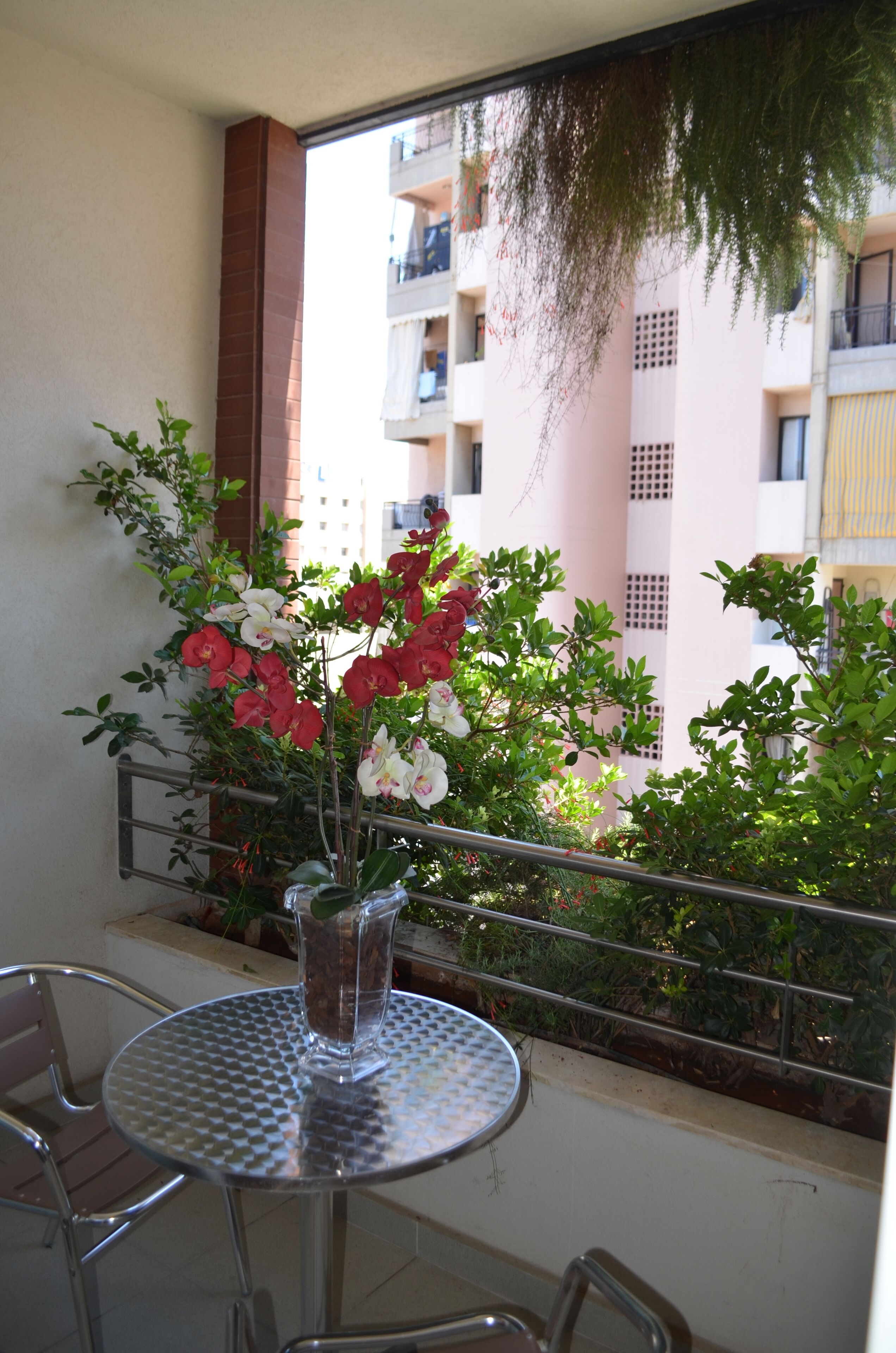 Un balcon avec une table et des chaises en métal, un pot de fleurs contenant des fleurs rouges et blanches, et une vue sur un immeuble doté de balcons.