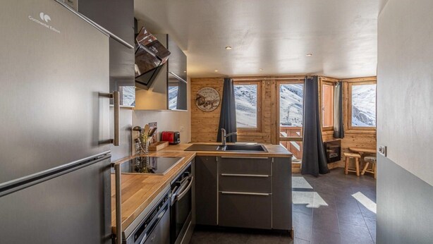 A modern kitchen with a stainless steel refrigerator, wooden countertops, and a built-in oven. There is a wooden dining area with a bench and a view of a snowy landscape through large windows.