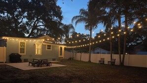 Un patio trasero con una casa pequeña, luces colgantes, una parrilla y mobiliario exterior.