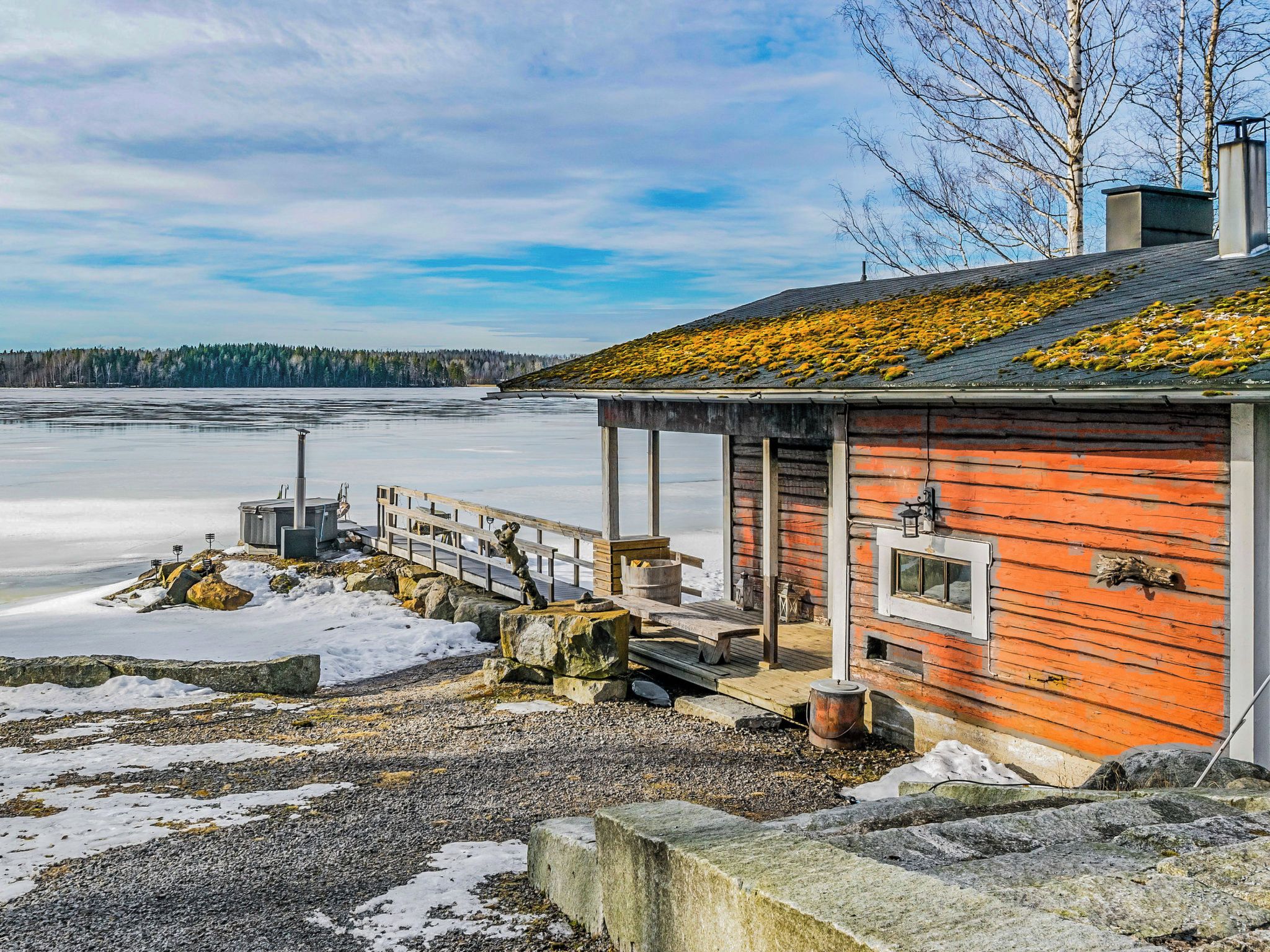 A wooden cabin with a dock extending into a frozen lake, surrounded by trees and a clear sky.