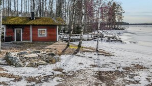 Une petite cabane rouge avec une porte noire, entourée d’arbres et bordée d’un lac gelé.