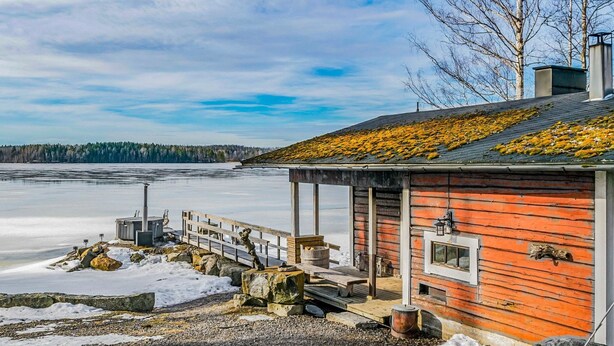 Une cabane en bois avec un quai s’avançant sur un lac gelé, entourée de rochers et de neige.