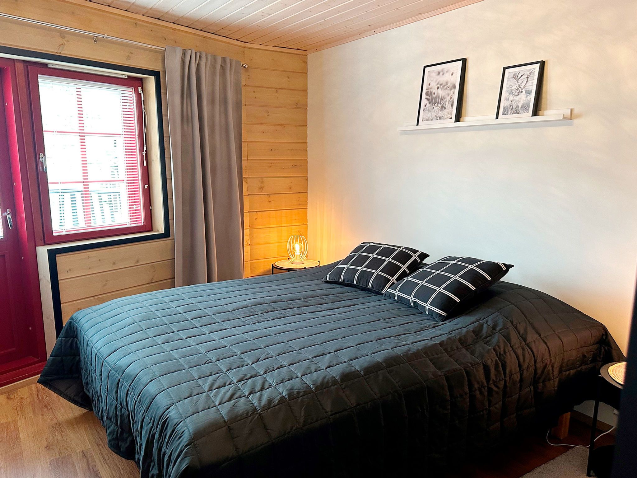 A bedroom with a bed, a window with blinds, a lamp, and two framed pictures on the wall.