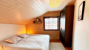A bedroom with a sloped ceiling, a bed with a patterned bedspread, a window with blinds, a bedside lamp, and a dark wardrobe.