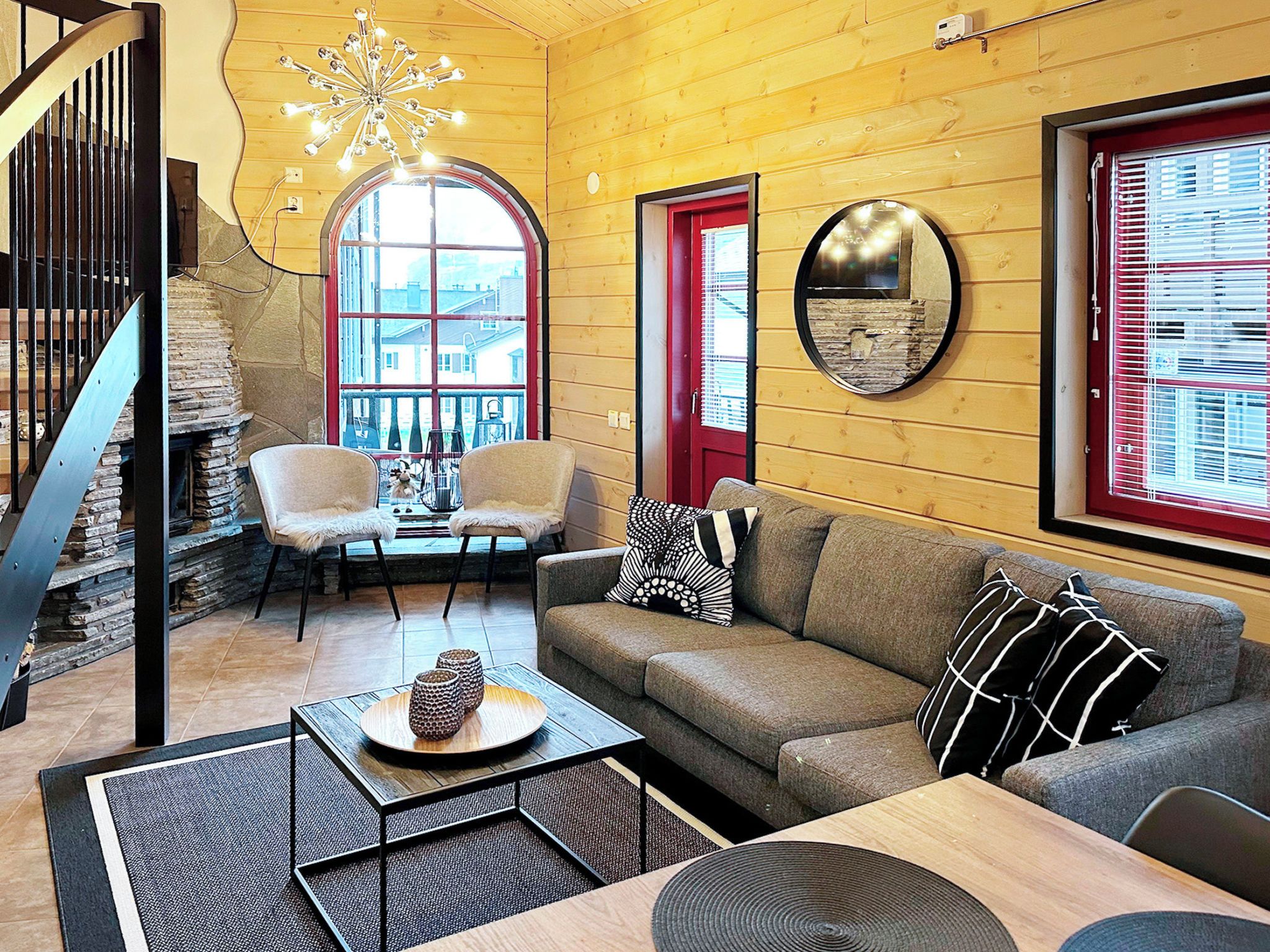 A cozy living room with a stone staircase, a wooden ceiling, a round mirror, and a red door.