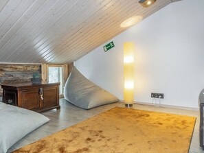 Una habitación con techo inclinado, un armario de madera, un sofá color beige, una alfombra amarilla y una lámpara de pie fijada a la pared.