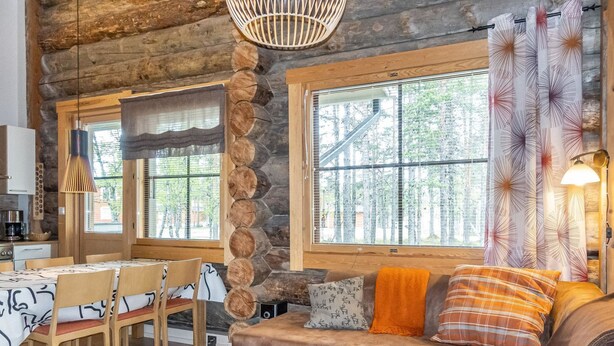 L’intérieur chaleureux d’un chalet en rondins comprend un coin repas, un canapé agrémenté de coussins à carreaux et de grandes fenêtres donnant sur la forêt.