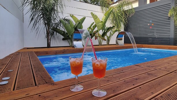 Een houten terras met een jacuzzi, twee glazen oranje drank en een waterfontein.