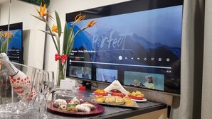 Een eethoek met een grote flatscreen-tv waarop een videoclip draait, een plateau met desserts en een decoratieve vaas met bloemen.