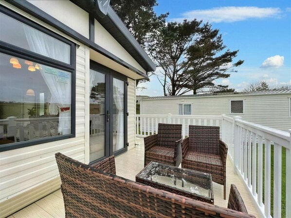 A covered outdoor seating area with wicker furniture and a glass table, adjacent to a white mobile home with a white fence. - Pottsville Cabin - Short Walk to Praa Sands Beach (Penzance)