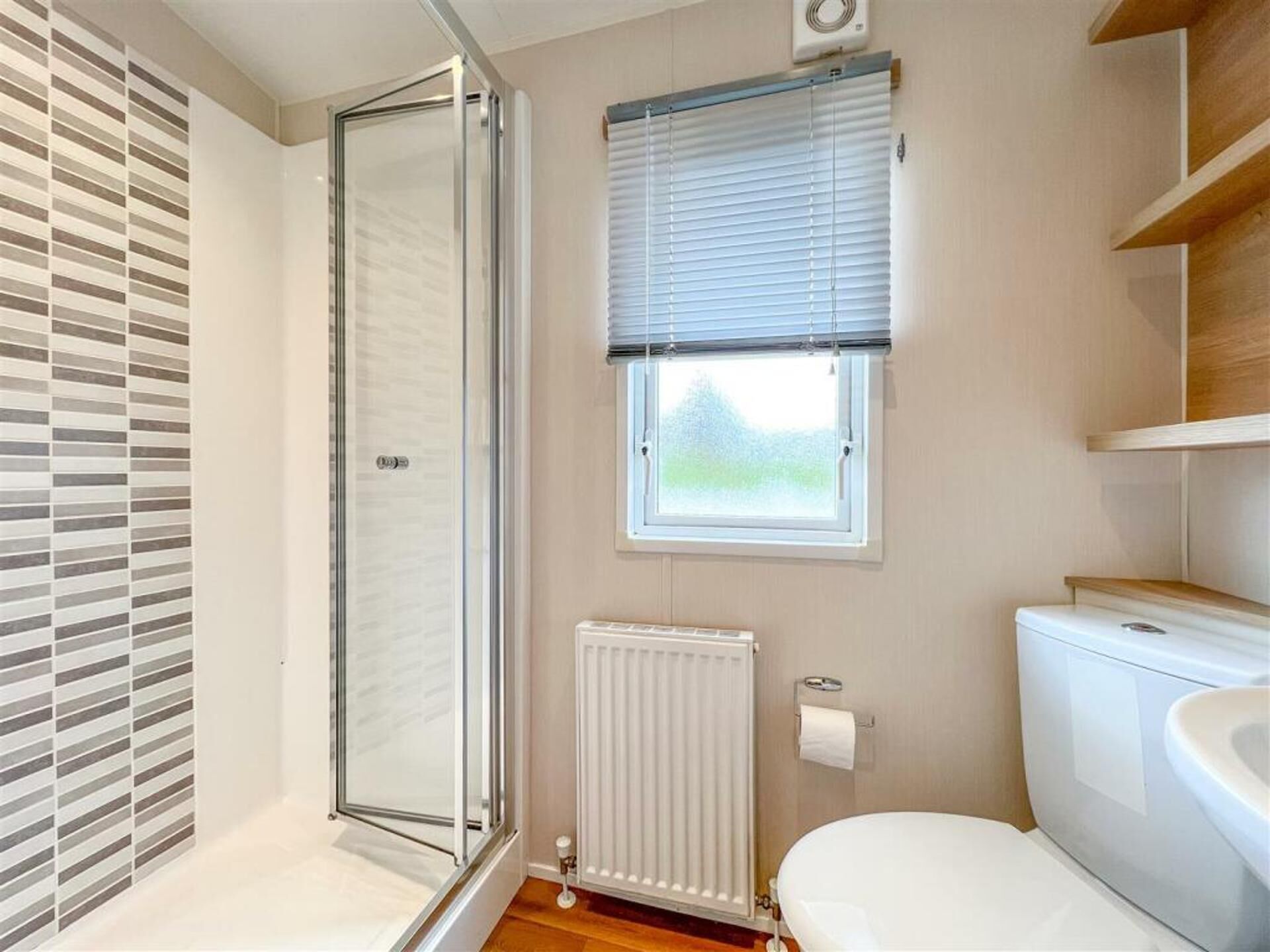 A bathroom with a shower, toilet, radiator, and a window with blinds.