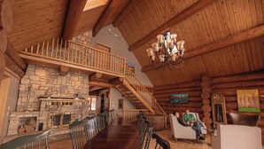 Un intérieur spacieux de chalet en bois, avec une longue table à manger, une cheminée, un escalier et un lustre.