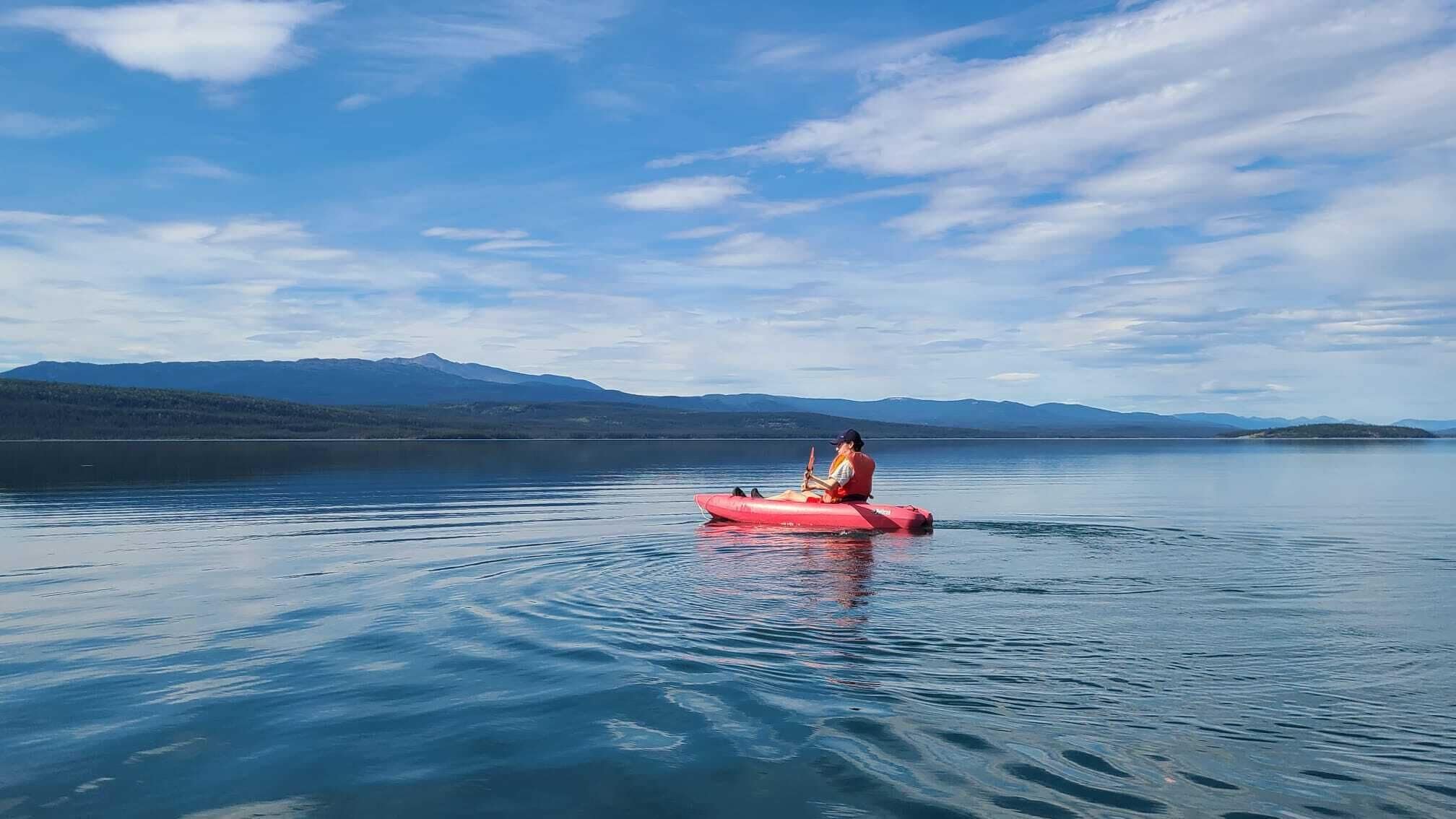 Une personne est assise sur un radeau gonflable rouge, sur un lac calme, avec des montagnes en arrière-plan.