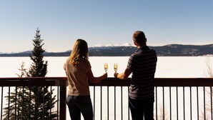 Deux personnes, chacune tenant un verre, se tiennent sur un balcon d’où l’on aperçoit un paysage enneigé.