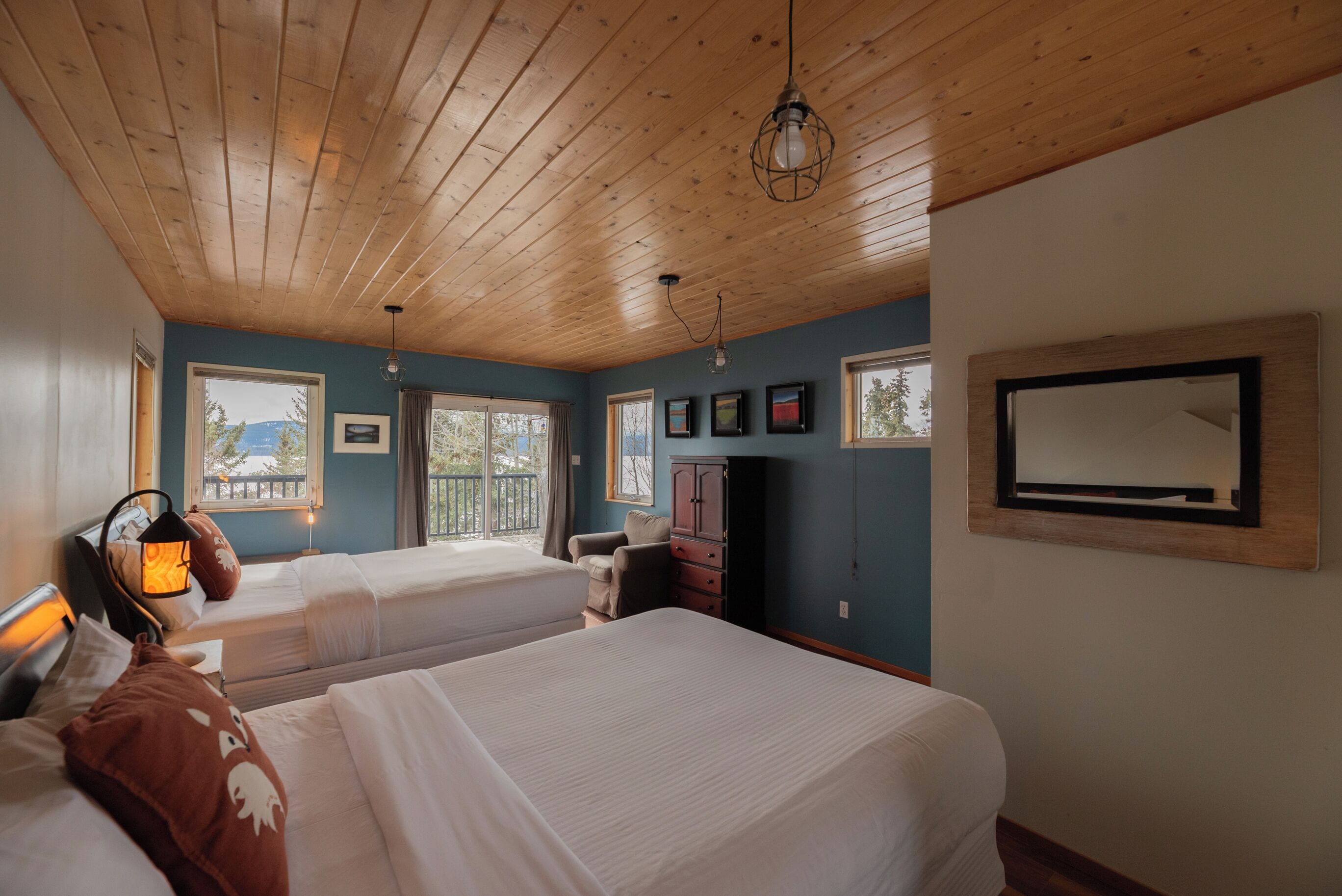 A room with two beds, wooden ceiling, large windows, and a TV mounted on the wall.