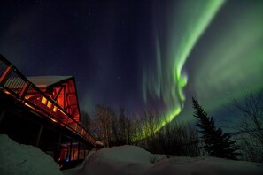A building with a red-lit structure, snow-covered ground, and a view of the Northern Lights. - Inn On The Lake (Marsh Lake)