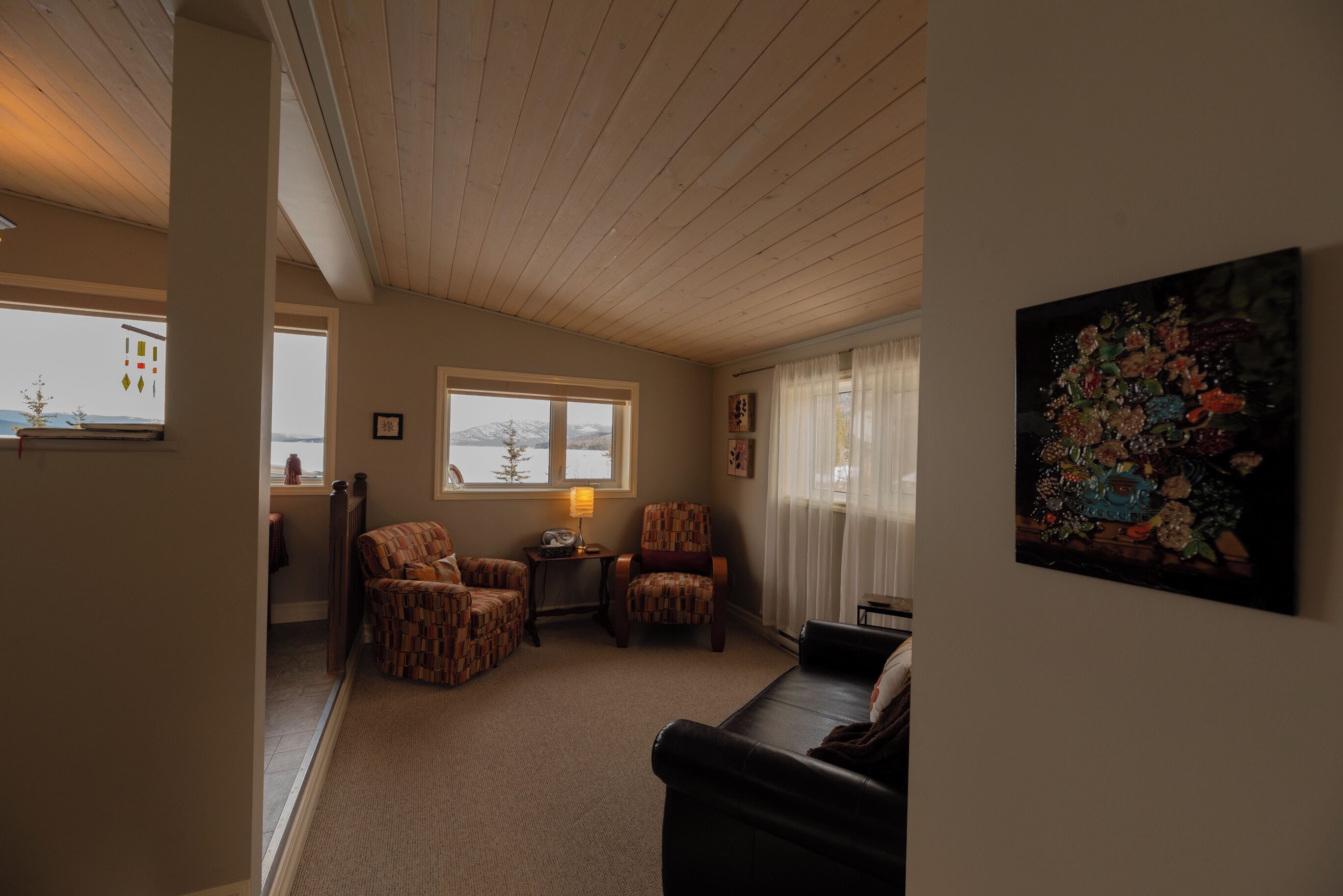 Een woonkamer met een houten plafond, een bank, twee gestreepte stoelen, een tafel met een lamp en een schilderij aan de muur.