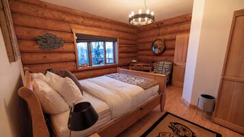 Un dormitorio de cabaña de troncos con cama de madera, lámpara de noche, ventana con vista a árboles, lámpara colgante y una pequeña mesa con sillas.