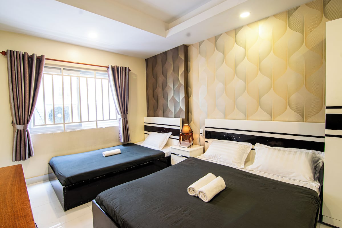 A hotel room with two beds, a window with curtains, a bedside table with a lamp, and a patterned wall.