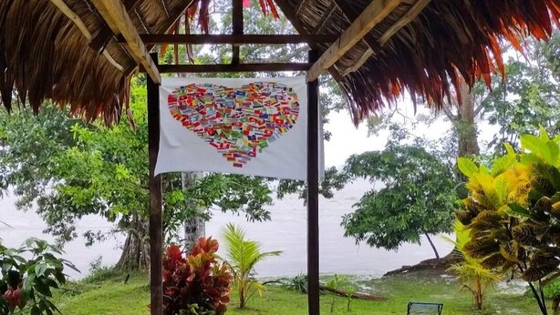A covered outdoor area with a thatched roof, a heart-shaped artwork hanging from the ceiling, and a grassy area with chairs and plants. - Iquitos (San Juan Bautista)