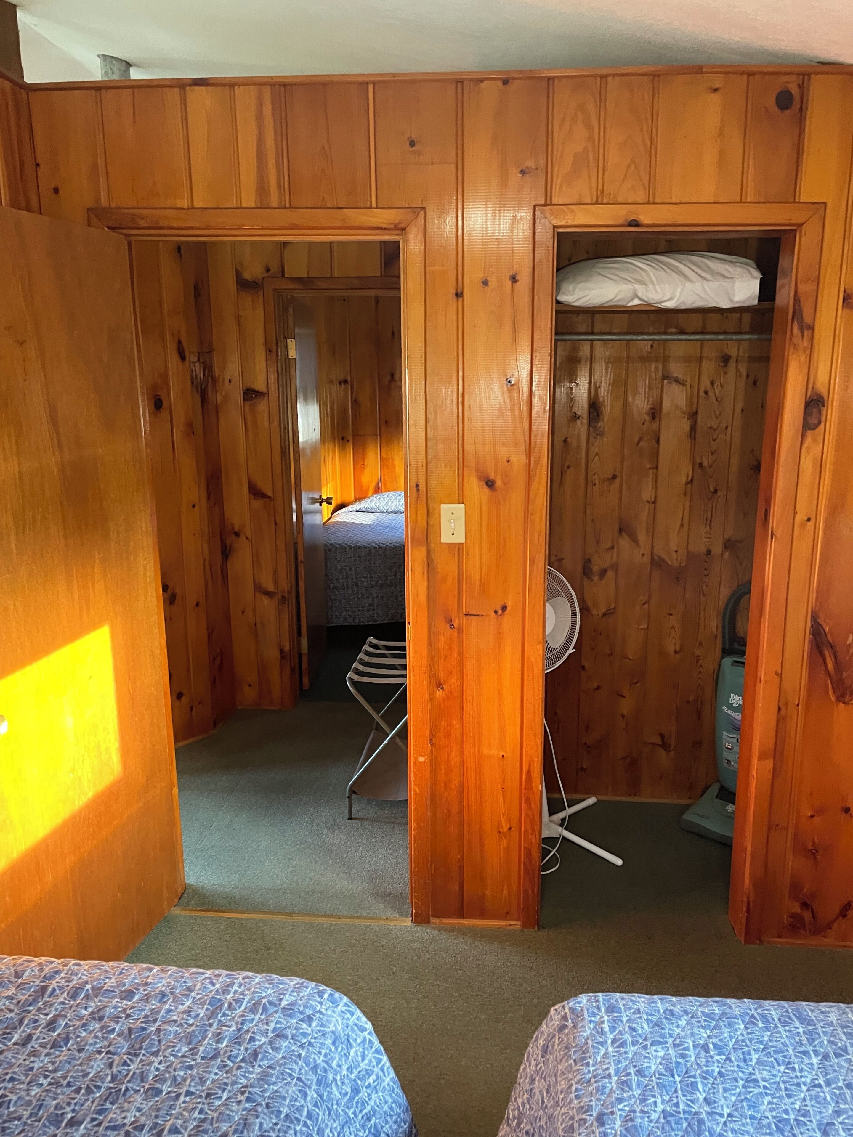 A room with wooden walls, a bed, a fan, and a vacuum cleaner.