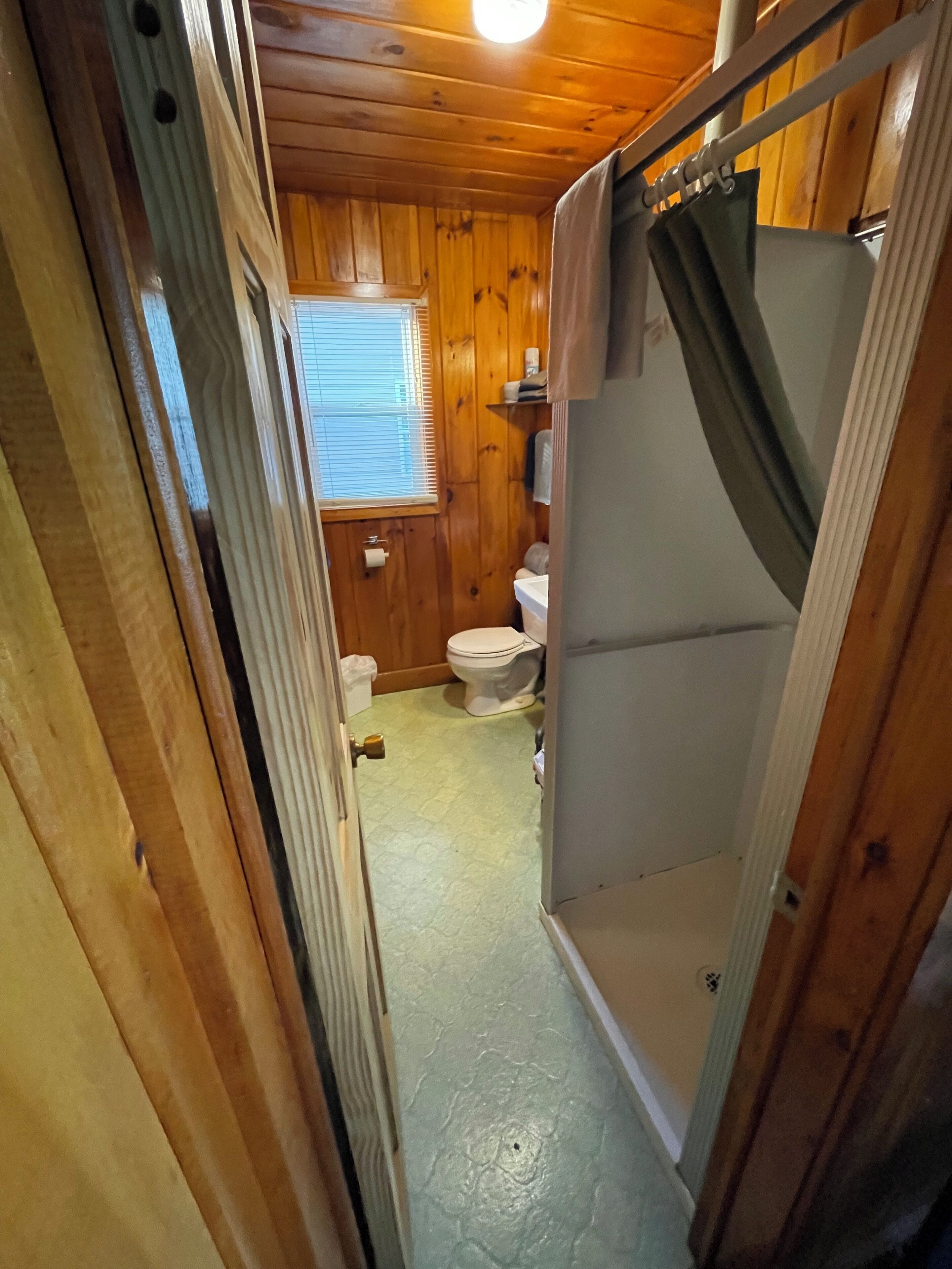 A narrow bathroom with wooden walls, a toilet, and a shower area.