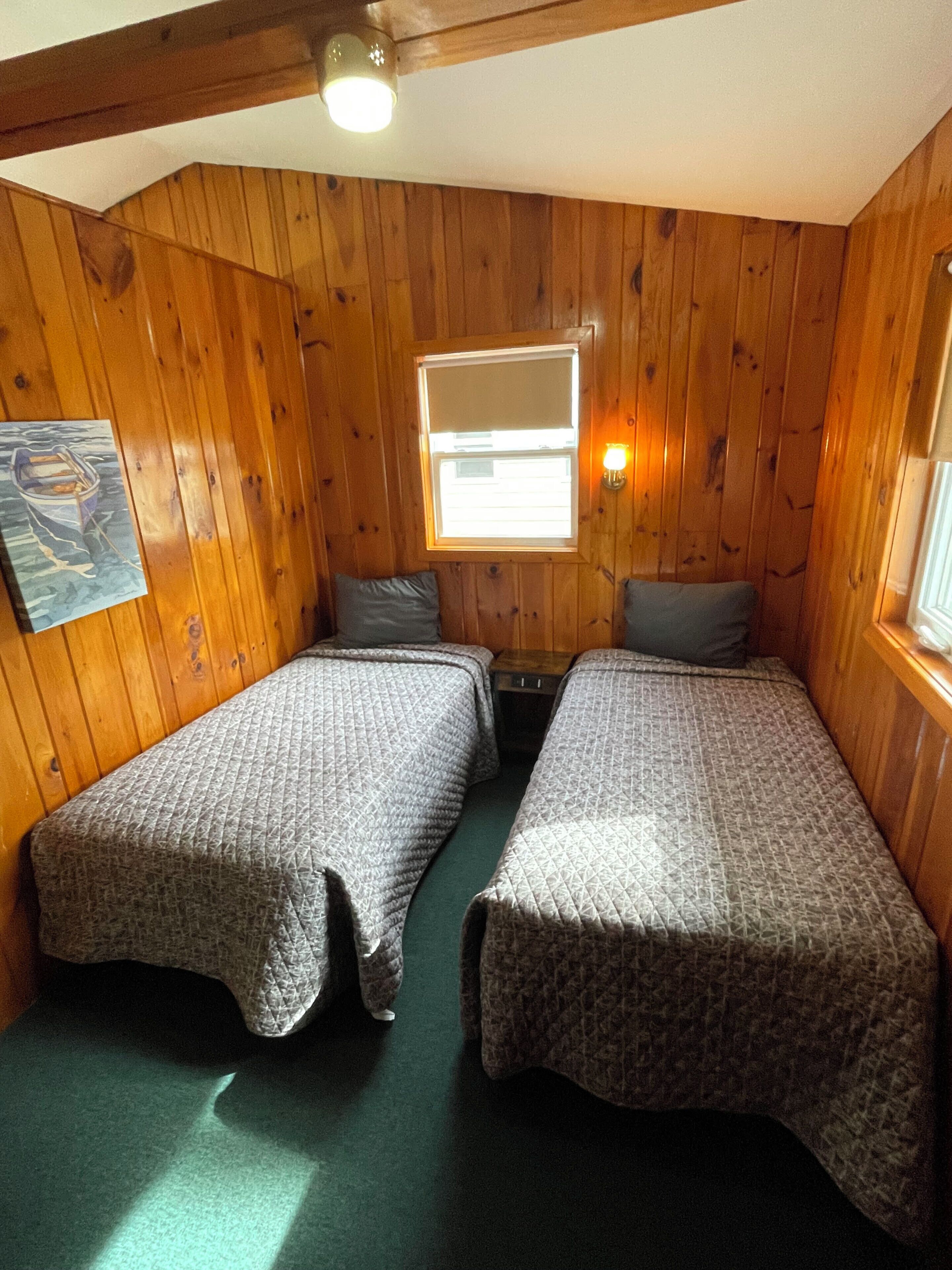 Una habitación con paredes de madera, dos camas con ropa de cama gris, una ventana con persianas, una lámpara fijada a la pared y un cuadro en la pared izquierda.