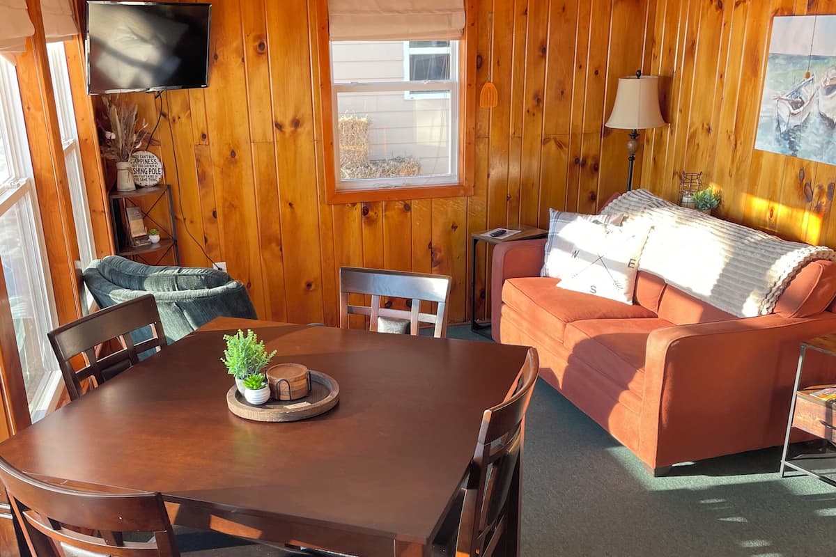 A wooden-paneled room with a dining table, chairs, a sofa, a TV, and a window.