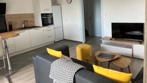 A modern kitchen with white cabinets, a black refrigerator, and a built-in oven. There is a dining area with a small table and chairs, and a living room with a sofa and a TV.