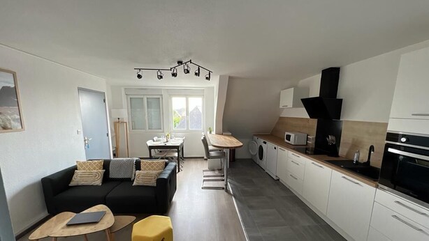 A modern kitchen with white cabinets, a black stove, and a wooden countertop. There is a dining area with a table and chairs, and a living room with a sofa and a yellow ottoman.