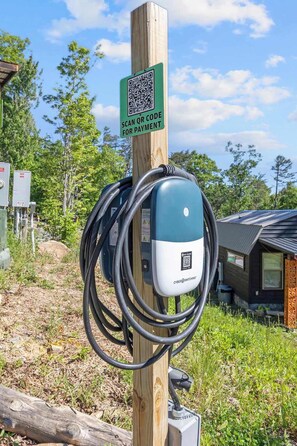 Un panneau avec un code QR pour effectuer le paiement, fixé sur un poteau en bois, une borne de recharge avec un câble, et un bâtiment en arrière-plan.