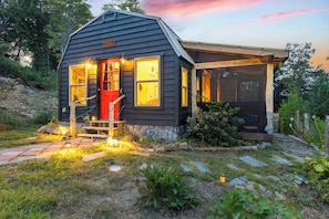 Une petite cabane aux couleurs sombres, dotée d’un porche et d’un espace extérieur abrité, et entourée d’un paysage naturel.
