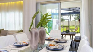Un espace repas avec une table en verre, des chaises à revêtement blanc et une vue sur l’extérieur grâce à de grandes portes-fenêtres coulissantes.