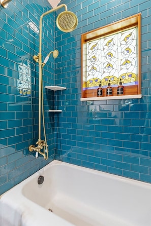 Une salle de bain avec une baignoire, une douche dotée d’un pommeau doré et un vitrail au-dessus du lavabo.