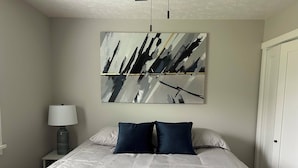Ein Schlafzimmer mit einem großen Bett, zwei Kissen, einem Nachttisch mit Lampe und einem an der Wand hängenden abstrakten Gemälde.