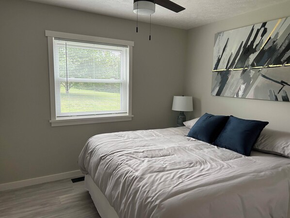 A bedroom with a bed, a window with blinds, a ceiling fan, a bedside table with a lamp, and a painting on the wall. - 3BR near skiing, snowshoeing, fishing, shopping & seasonal festivals downtown. (Traverse City)