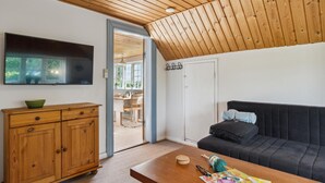 Une pièce chaleureuse avec un plafond en bois, un téléviseur à écran plat fixé au mur, un meuble en bois, un canapé noir avec des coussins et une petite table avec du matériel de tricot.