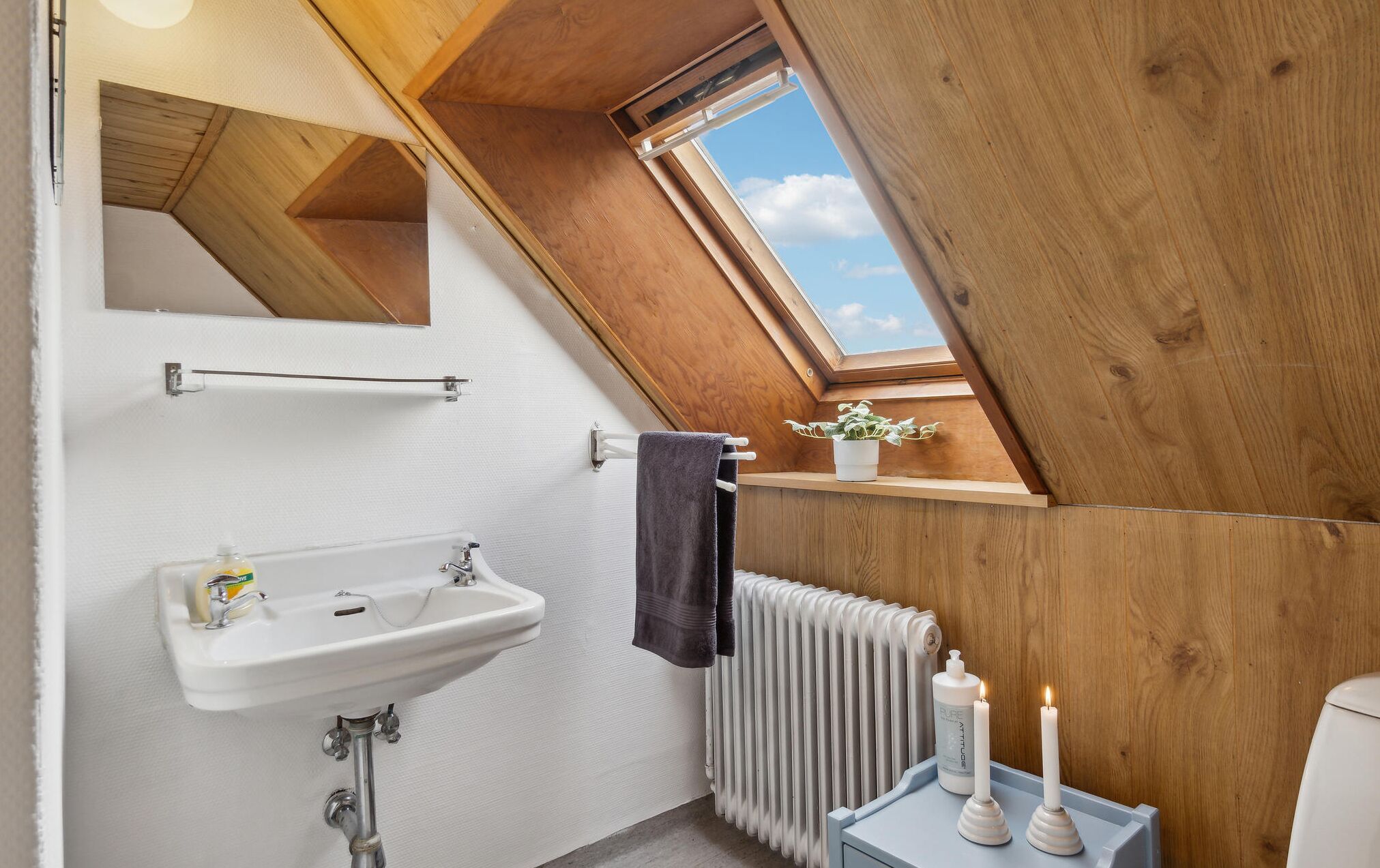 A small, cozy bathroom with a skylight, a white sink, a towel rack, a radiator, and a small shelf with a plant and candles.
