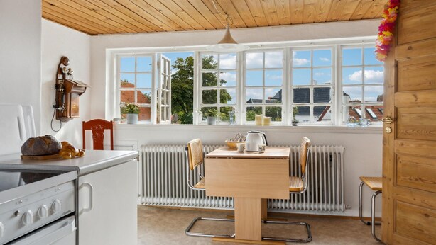 Une cuisine avec un plafond en bois, des murs blancs et une grande fenêtre à carreaux. On y trouve une cuisinière, une table avec des chaises et un radiateur.