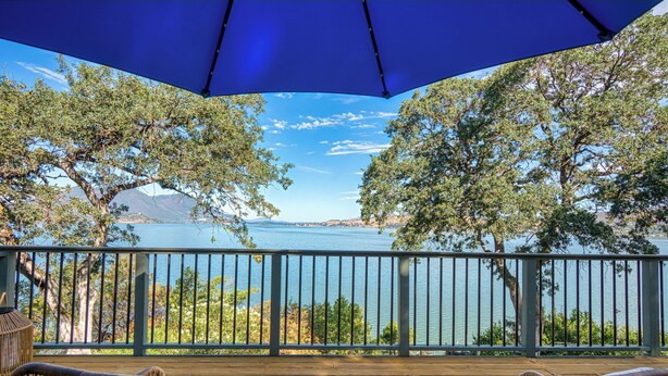 A wooden deck with wicker chairs and a blue umbrella, overlooking a lake with trees and mountains in the background. - Views! Private! Tranquil! Lakefront! Forest! Sunsets! Acreage! Docks! (Lower Lake)